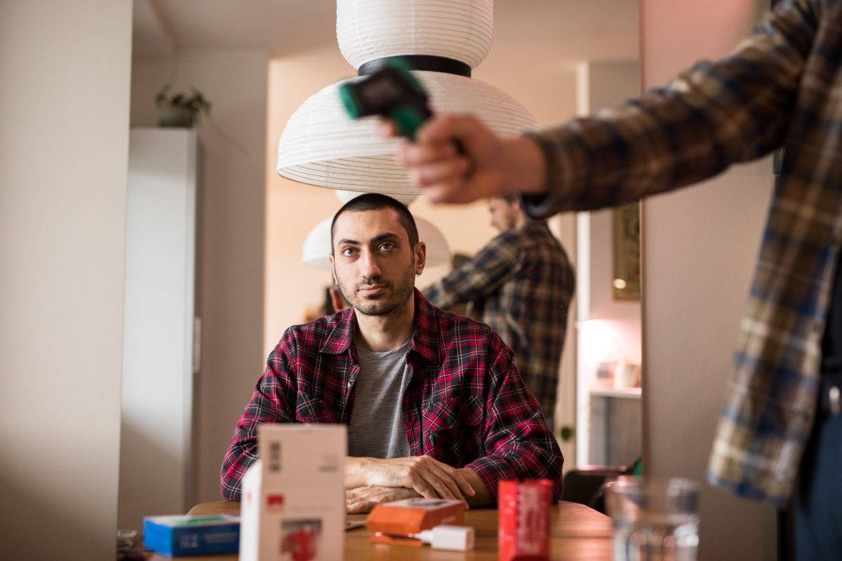 Henkilö istuu pöydän ääressä ja katsoo, kun toinen henkilö mittaa lämpötilaa lasermitalla.